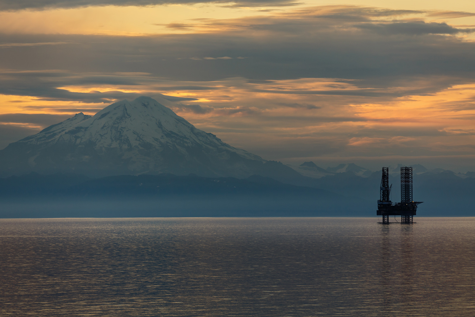 Oil and gas platform in Cook Inlet with snow covered mountain in background at sunset.